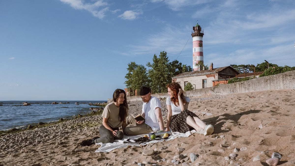 Friends enjoying a beach picnic with a lighthouse backdrop under a blue sky.